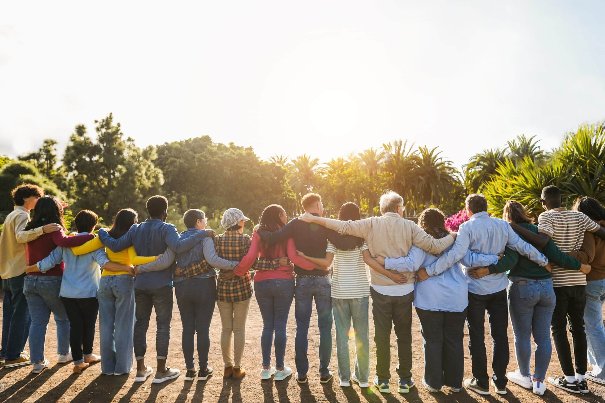 A group of people stand arm in arm facing a sunlit tree line.