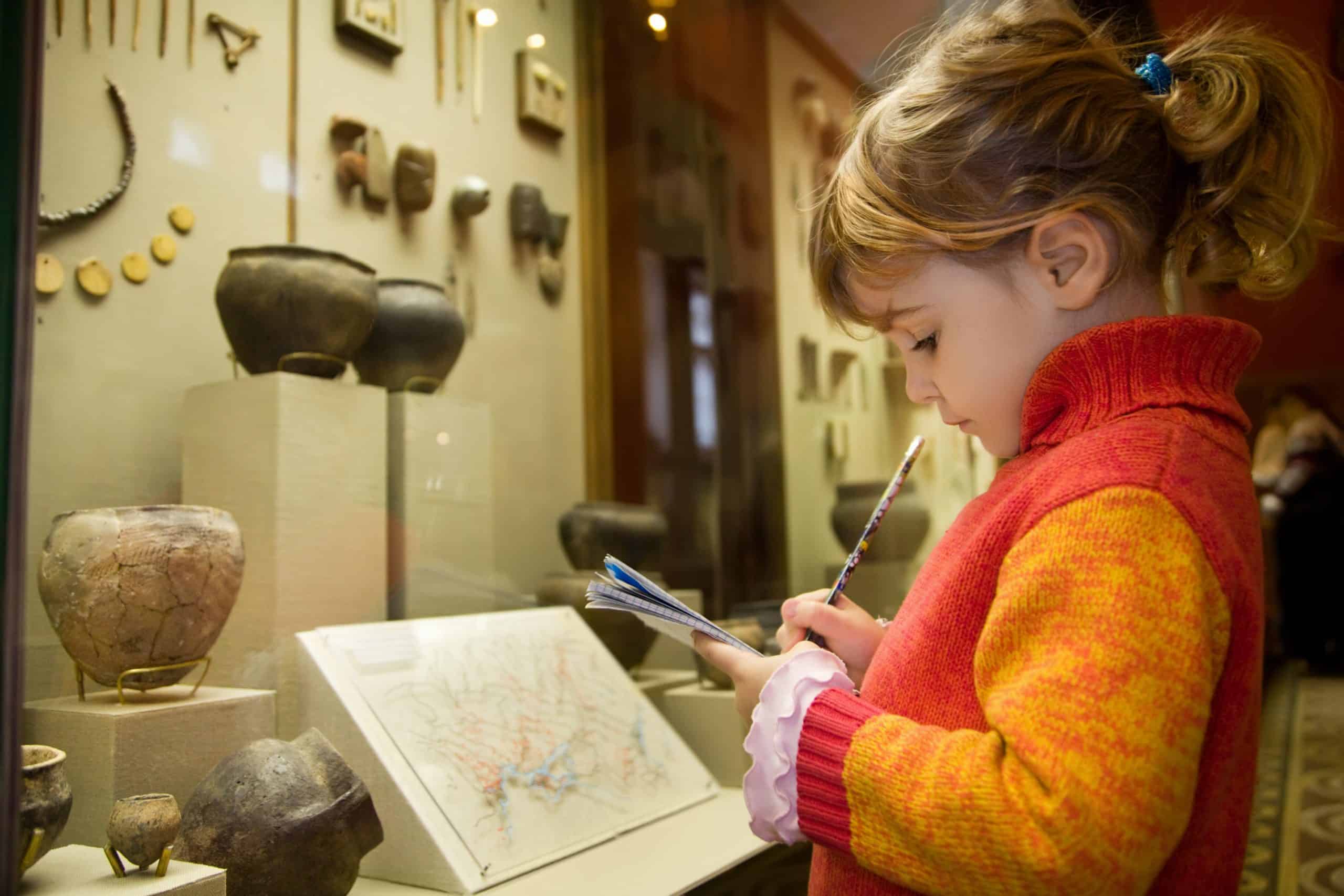 A young girl writes notes in a museum, standing beside a display case of pottery and artifacts.